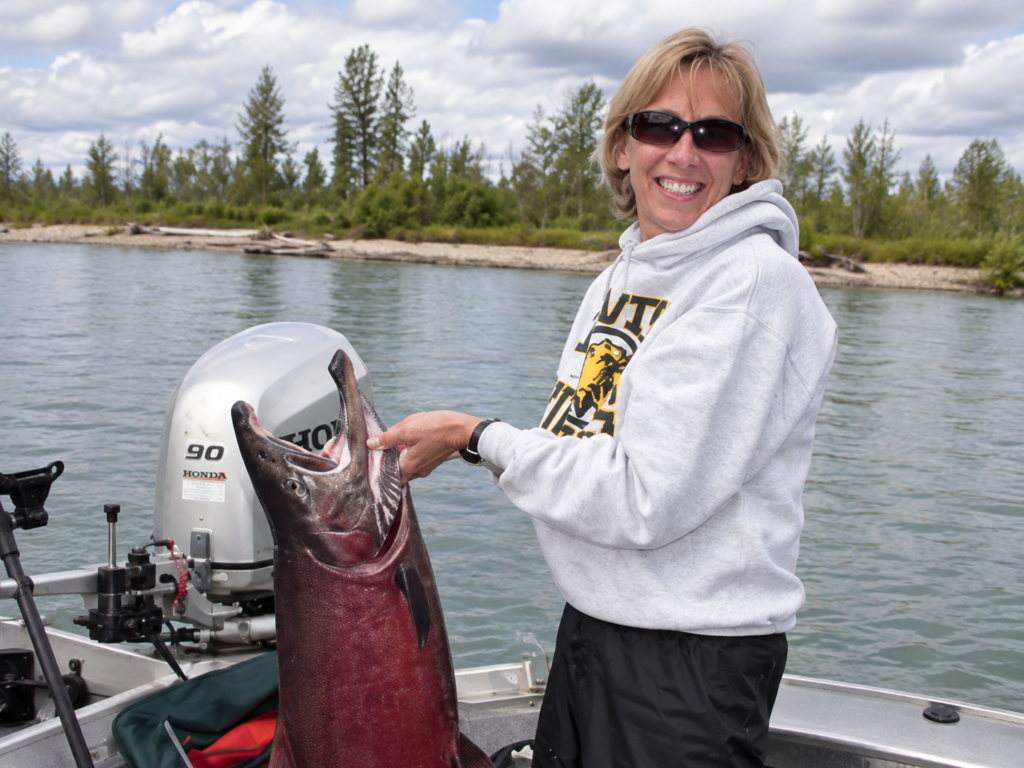Woman wearing sunglasses and a hoodie smiles while holding a large fish on a boat in a lake, with trees and clouds in the background.