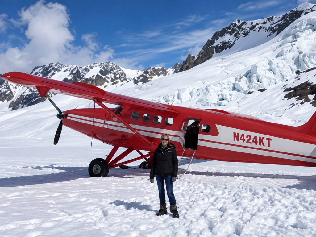 Person standing on snow in front of a red propeller plane with tail number N424KT, surrounded by snowy mountains and a glacier under a blue sky.