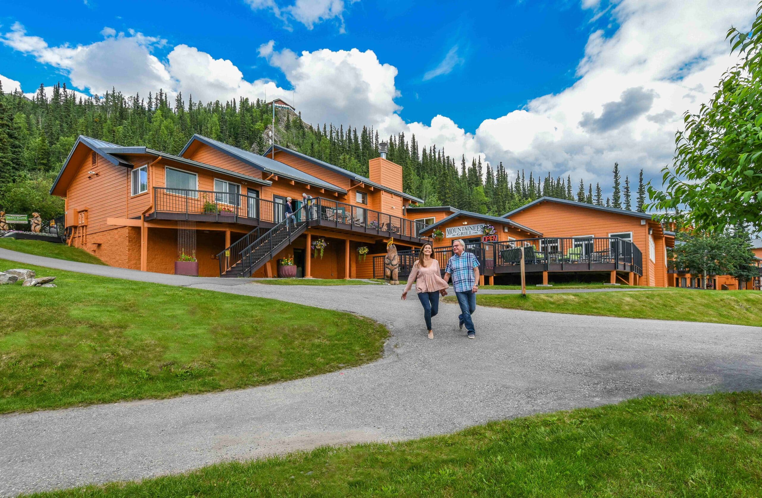 Two people walk on a paved path in front of several orange wooden lodges surrounded by green grass, trees, and a partly cloudy sky.
