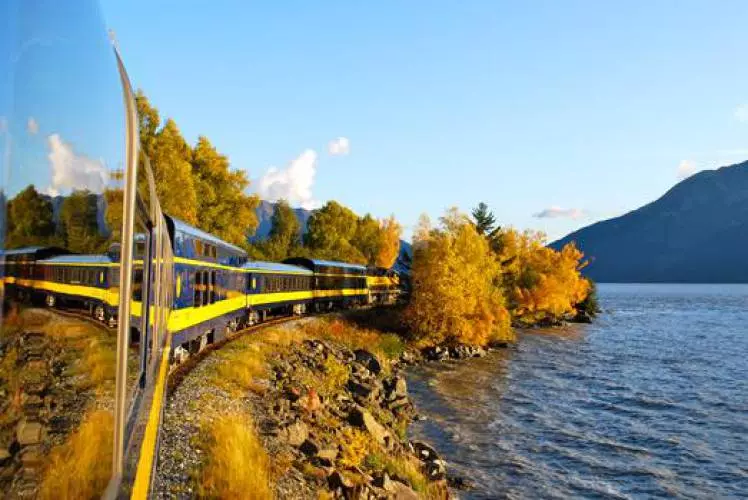A blue and yellow train travels along a lakeshore lined with autumn trees, with mountains and clear blue sky in the background.