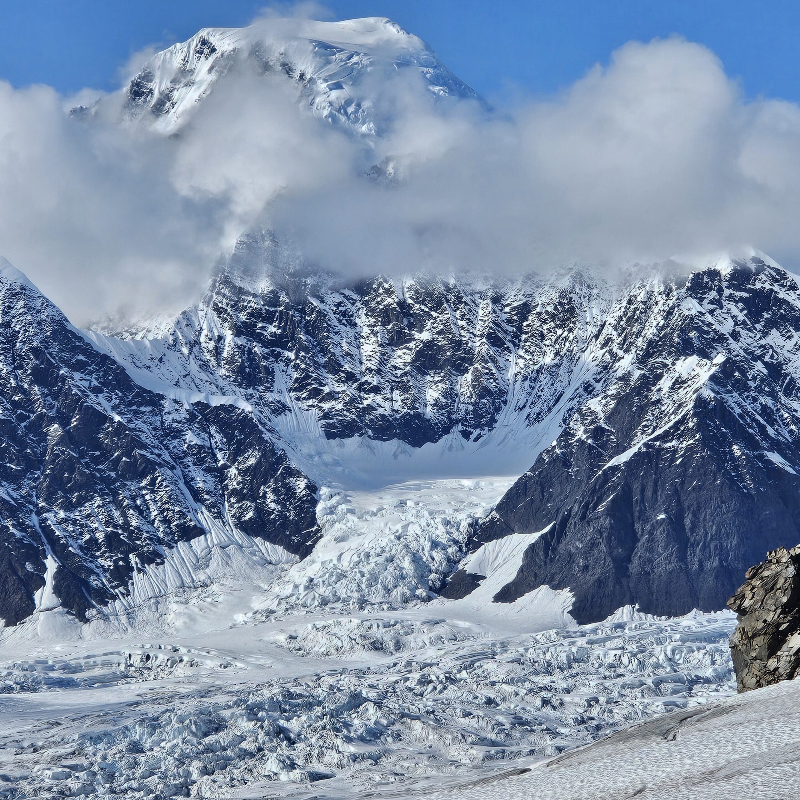 Snow-covered mountain with a glacier at its base, partially shrouded by clouds under a clear blue sky.