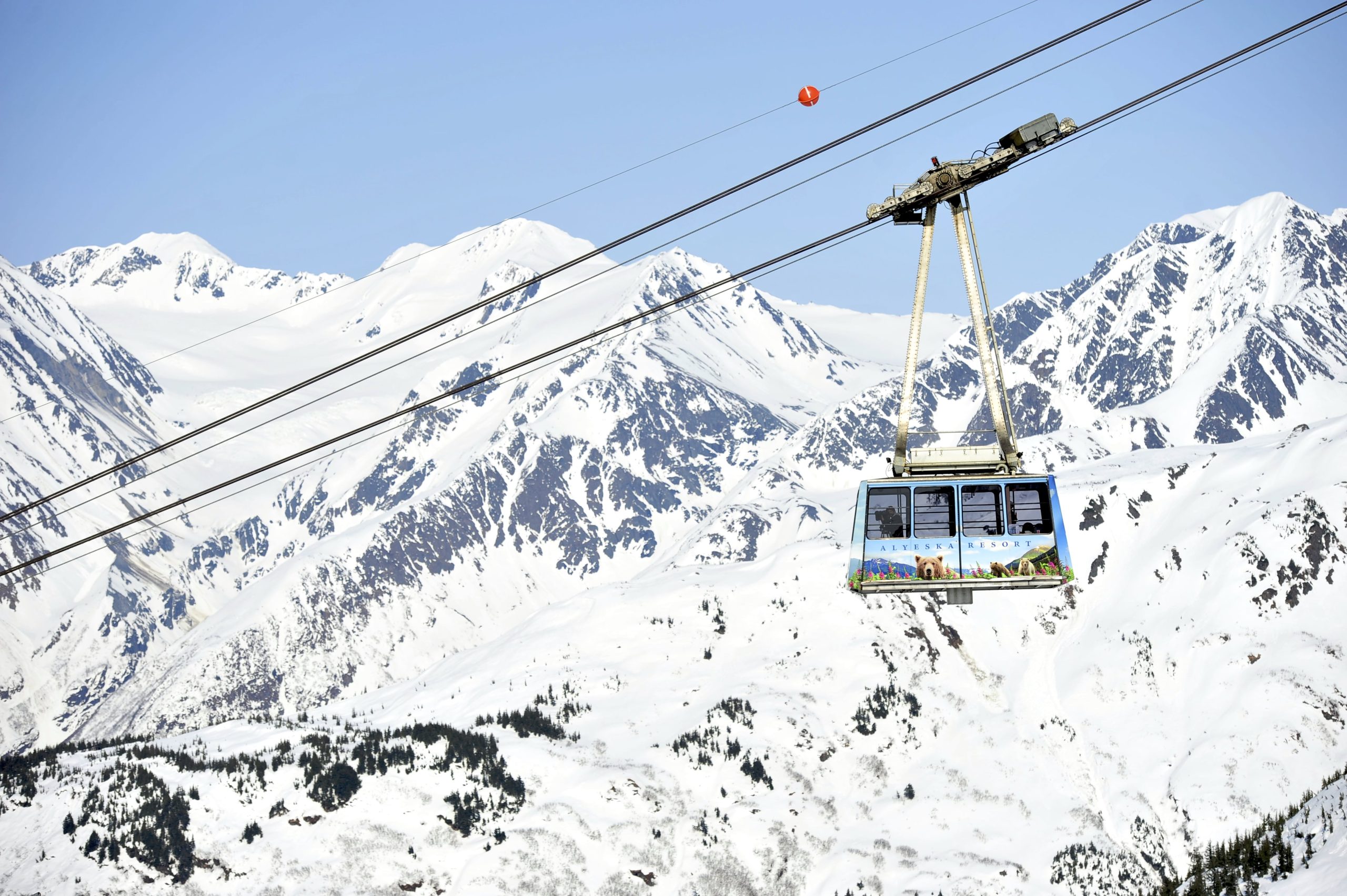 A cable car travels on suspended cables over snowy mountains under a clear sky.