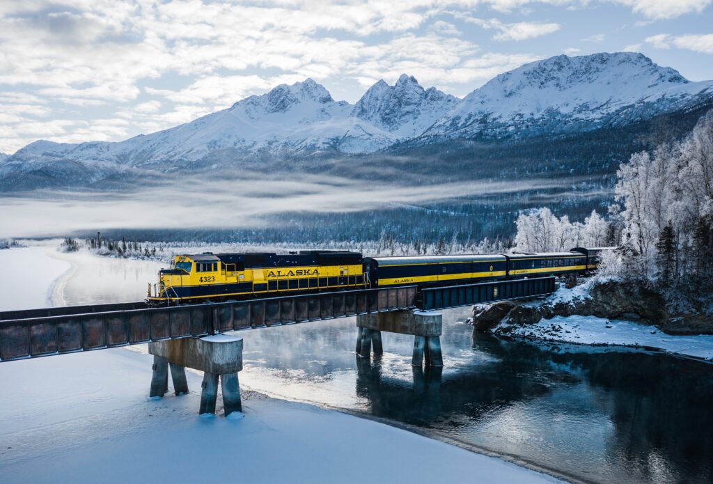 A yellow Alaska Railroad train crosses a bridge over a partially frozen river, with snow-covered mountains and trees in the background.