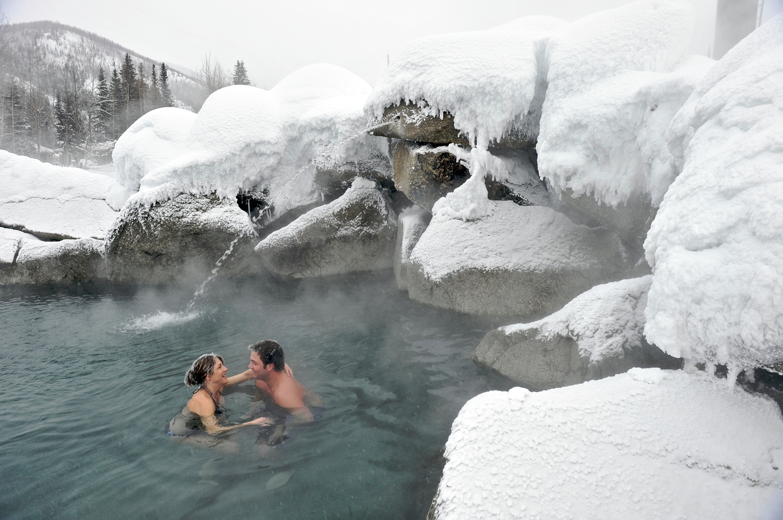 Two people relax in a steaming hot spring surrounded by snow-covered rocks and trees in a winter landscape.
