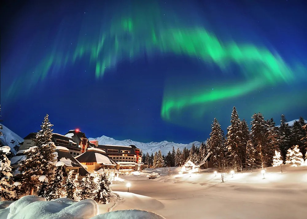 Snow-covered buildings and pine trees under a night sky with vivid green northern lights and distant mountain peaks.