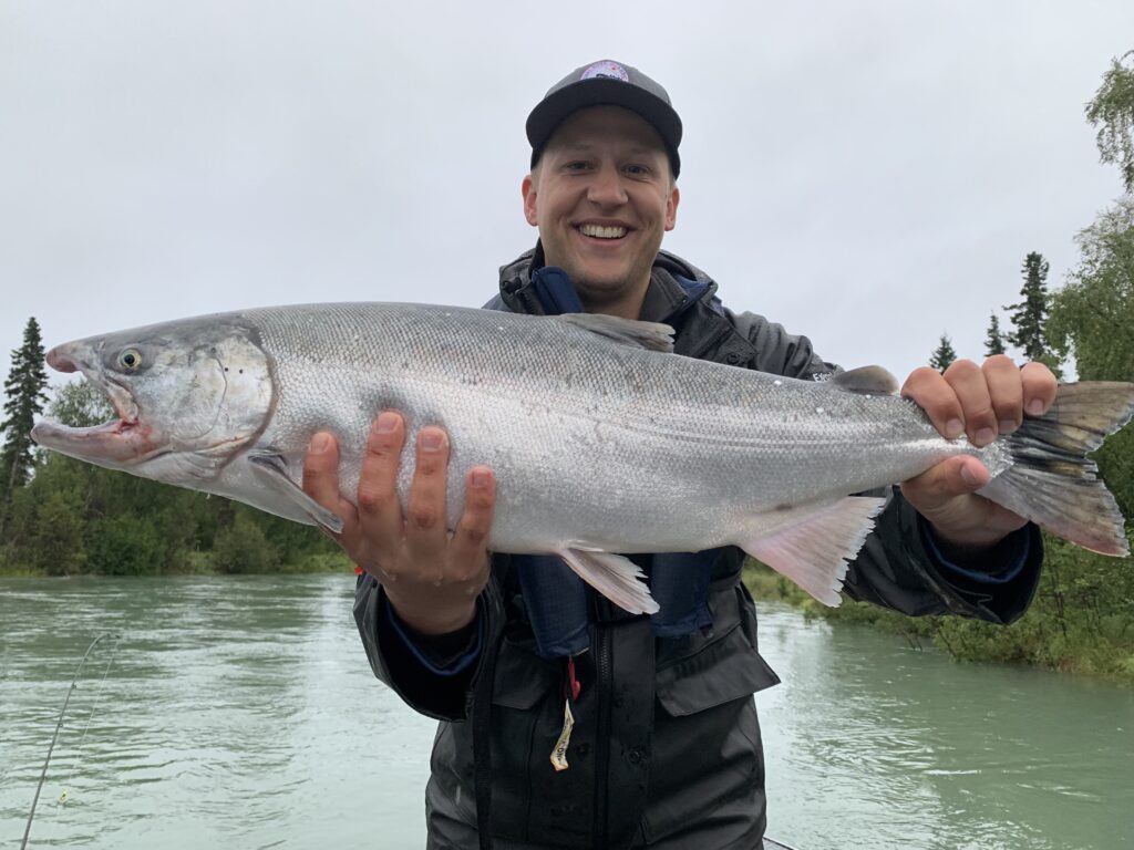 Person in outdoor gear holds a large silver fish with both hands near a river, smiling at the camera. Trees and overcast sky are visible in the background.