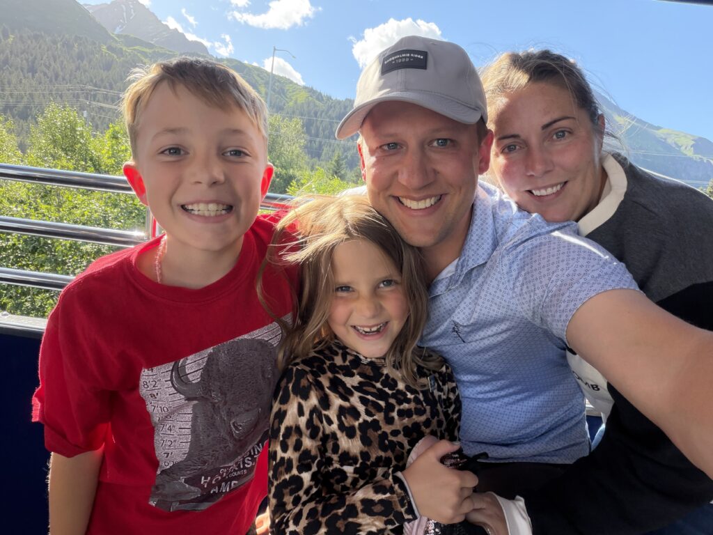 A smiling family of four takes a selfie outdoors on a sunny day, with trees, mountains, and blue sky in the background.