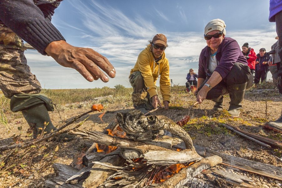 Three people crouch around a small campfire in a field, roasting sausages on sticks.