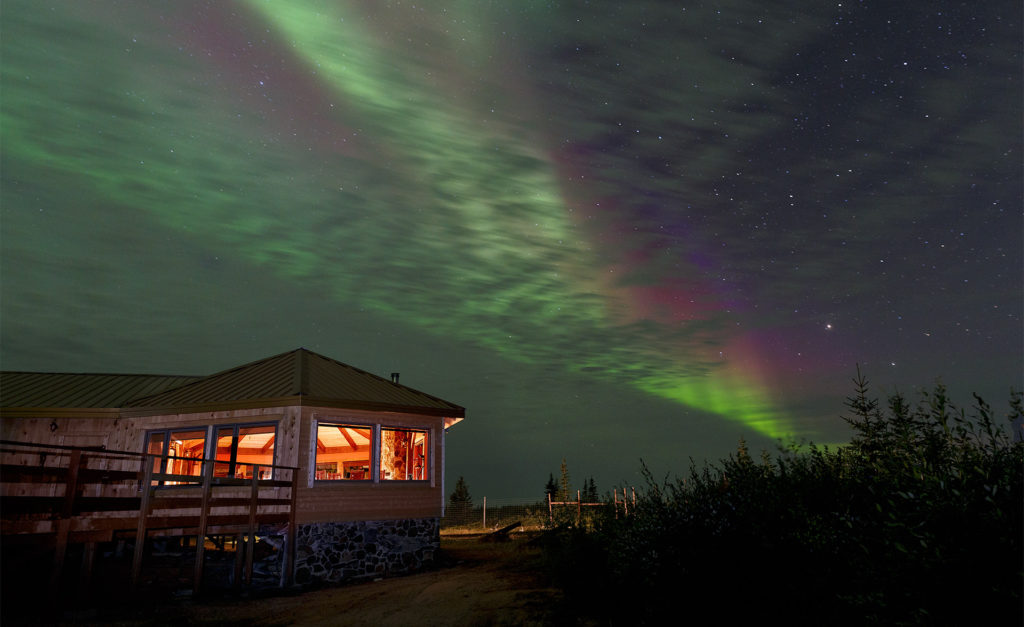 A brightly lit cabin at night under a sky filled with the aurora borealis.
