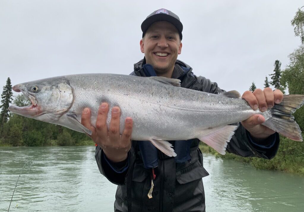 Person in outdoor gear holds a large silver fish with both hands near a river, smiling at the camera. Trees and overcast sky are visible in the background.