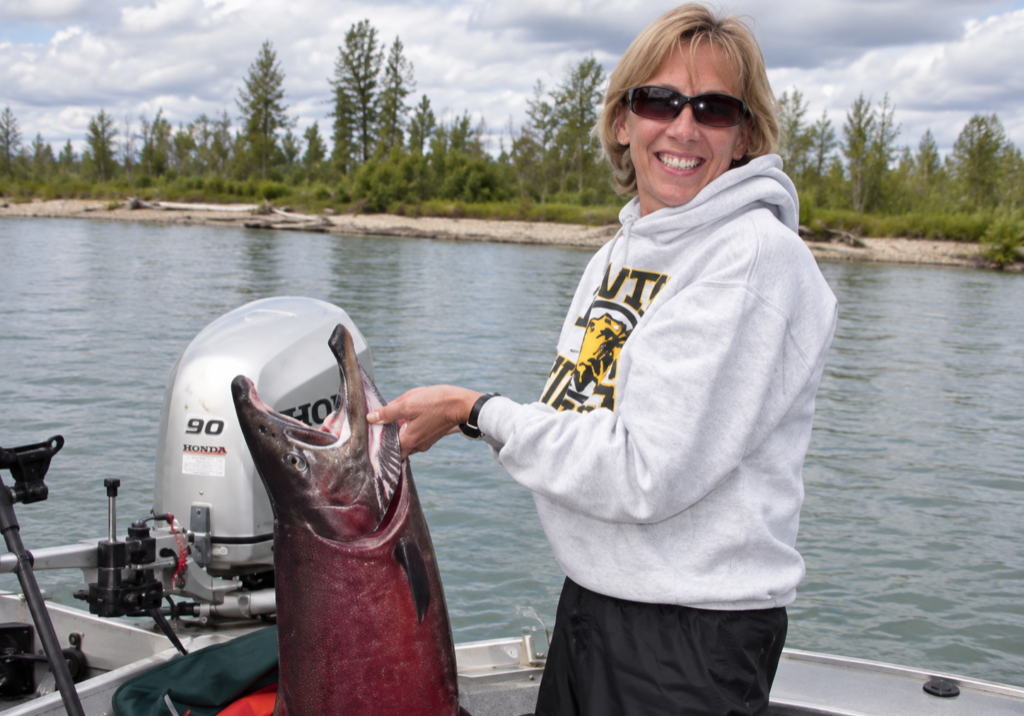 Woman wearing sunglasses and a hoodie smiles while holding a large fish on a boat in a lake, with trees and clouds in the background.