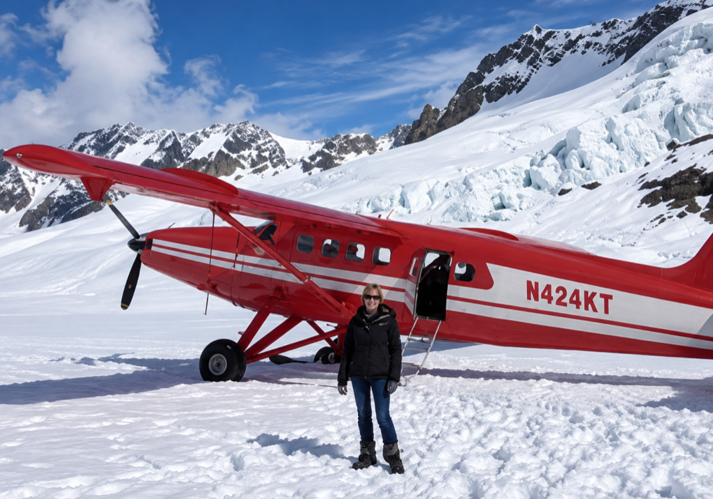 Person standing on snow in front of a red propeller plane with tail number N424KT, surrounded by snowy mountains and a glacier under a blue sky.