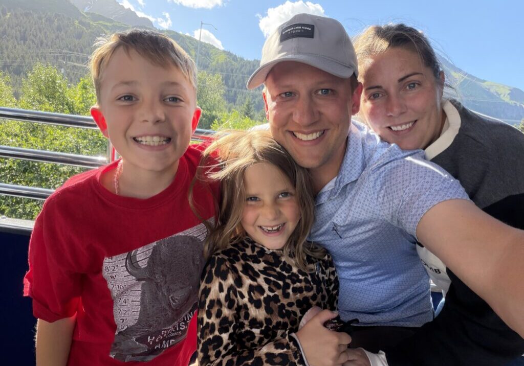 A smiling family of four takes a selfie outdoors on a sunny day, with trees, mountains, and blue sky in the background.